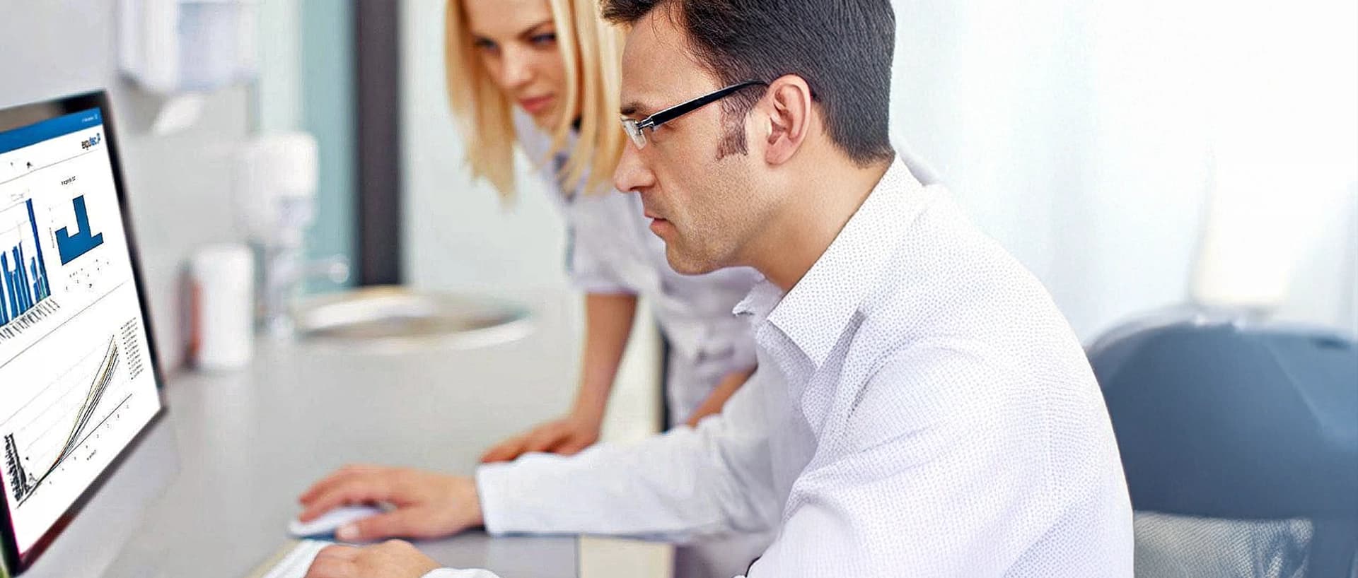 A man and woman in lab coats examining a computer screen displaying pharmaceutical software.
