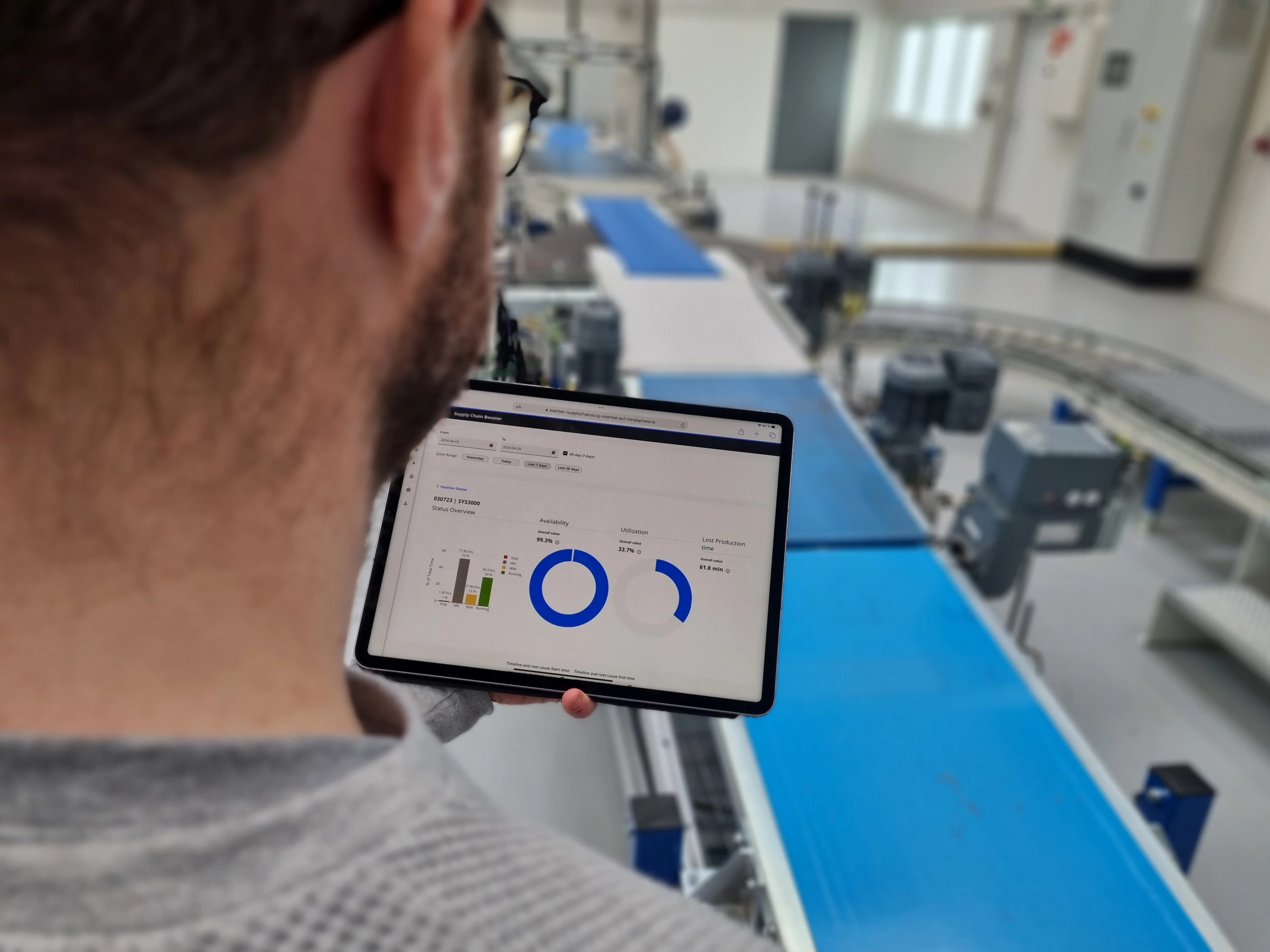 Person holding a tablet with maintenance analytics dashboard in front of an automated conveyor system in a warehouse.
