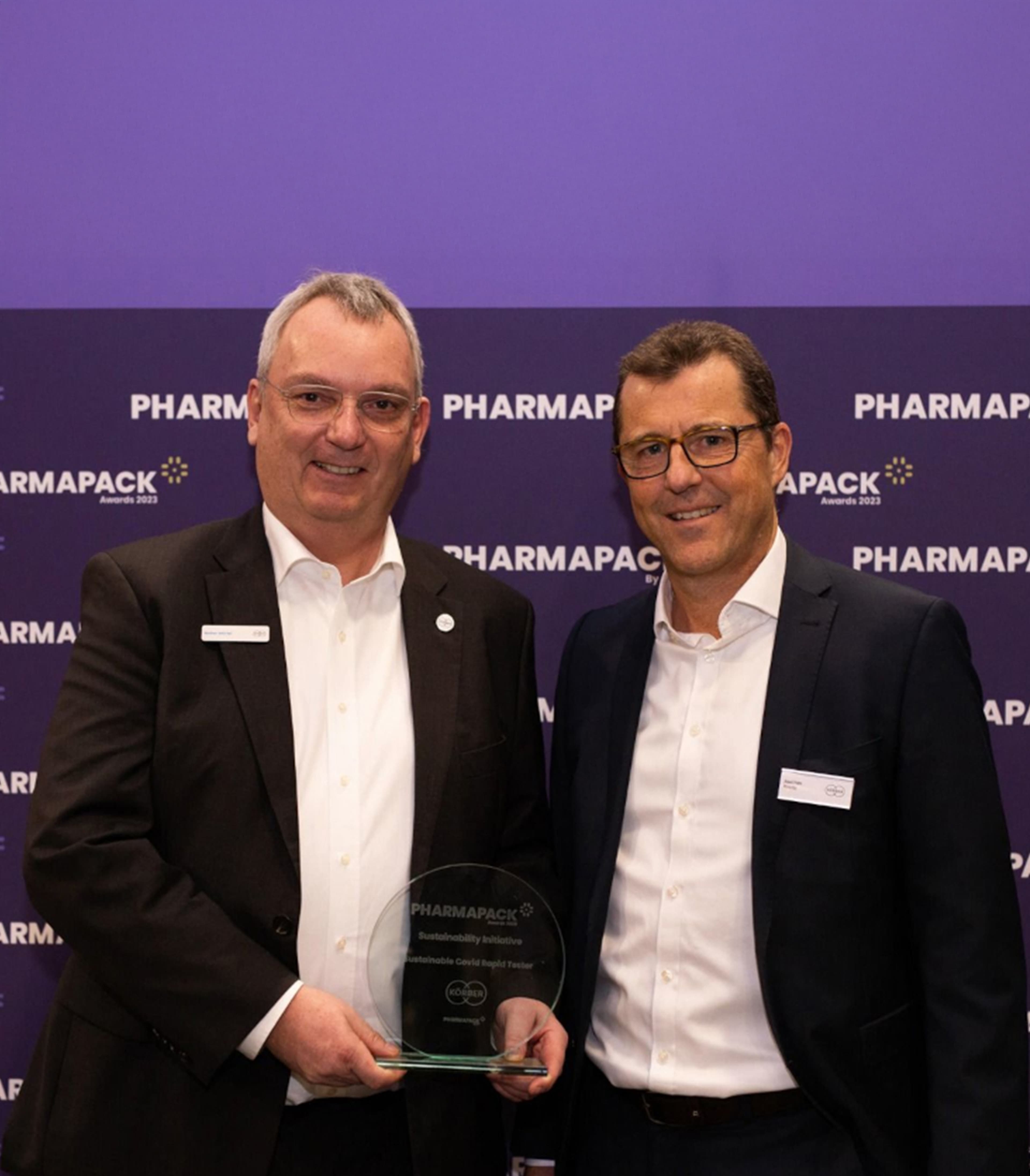 Two men smiling at the camera standing in front of a Pharmapack branded wall