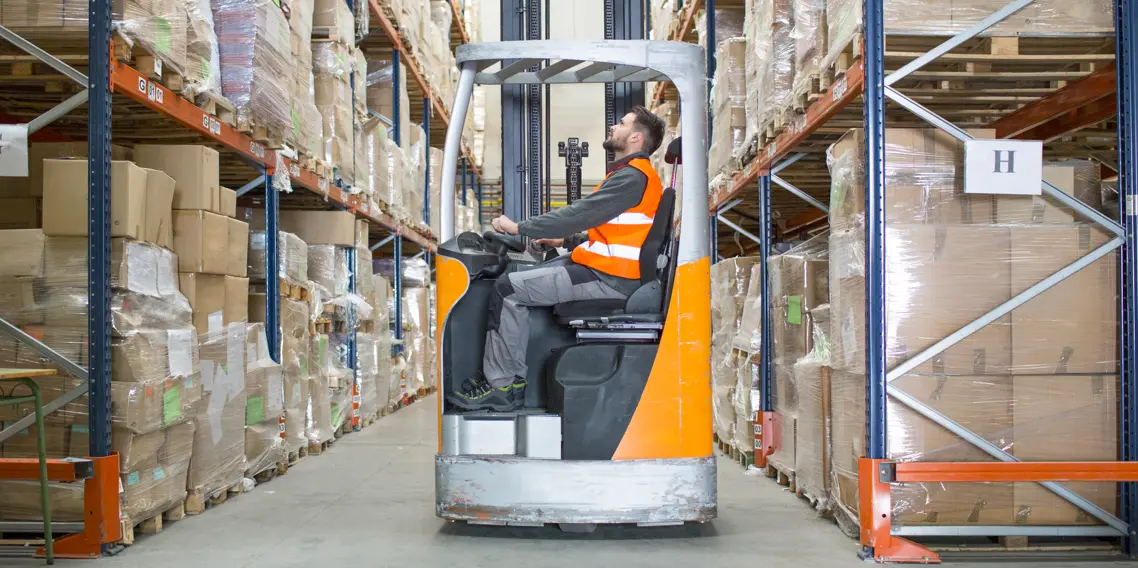 A man operating a forklift inside a warehouse, surrounded by shelves and stacked boxes.