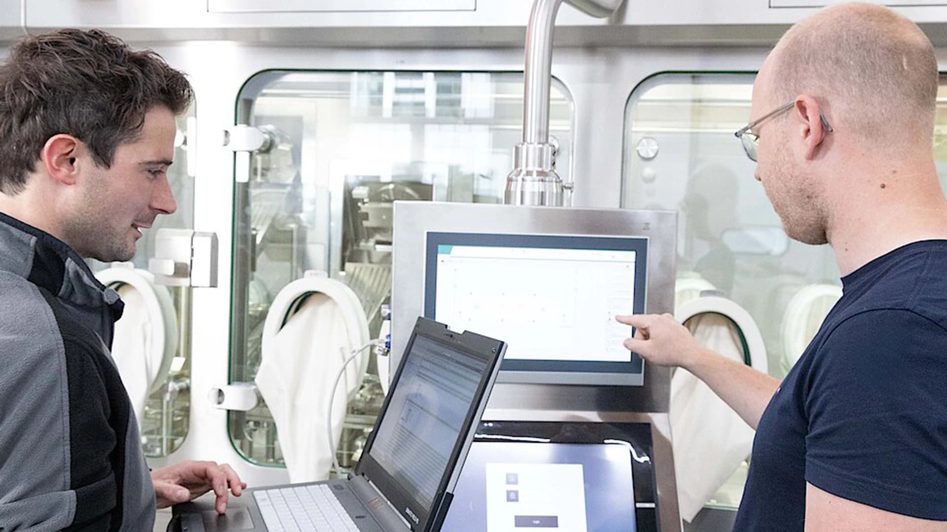 Two men collaborating on a laptop in a pharmaceutical factory setting.