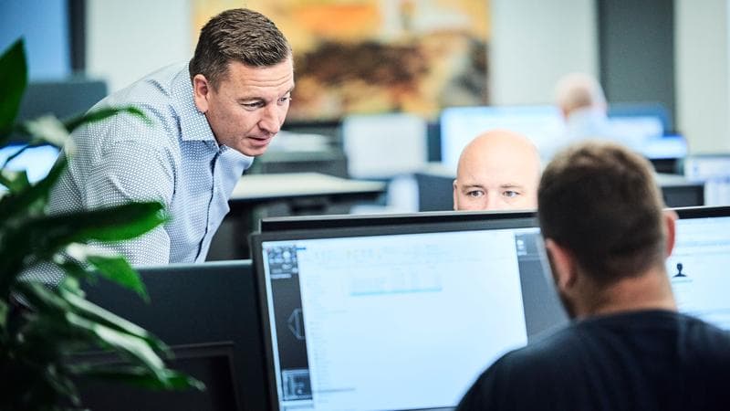 Zwei Männer arbeiten an Computern, während einer von ihnen sie beaufsichtigt.