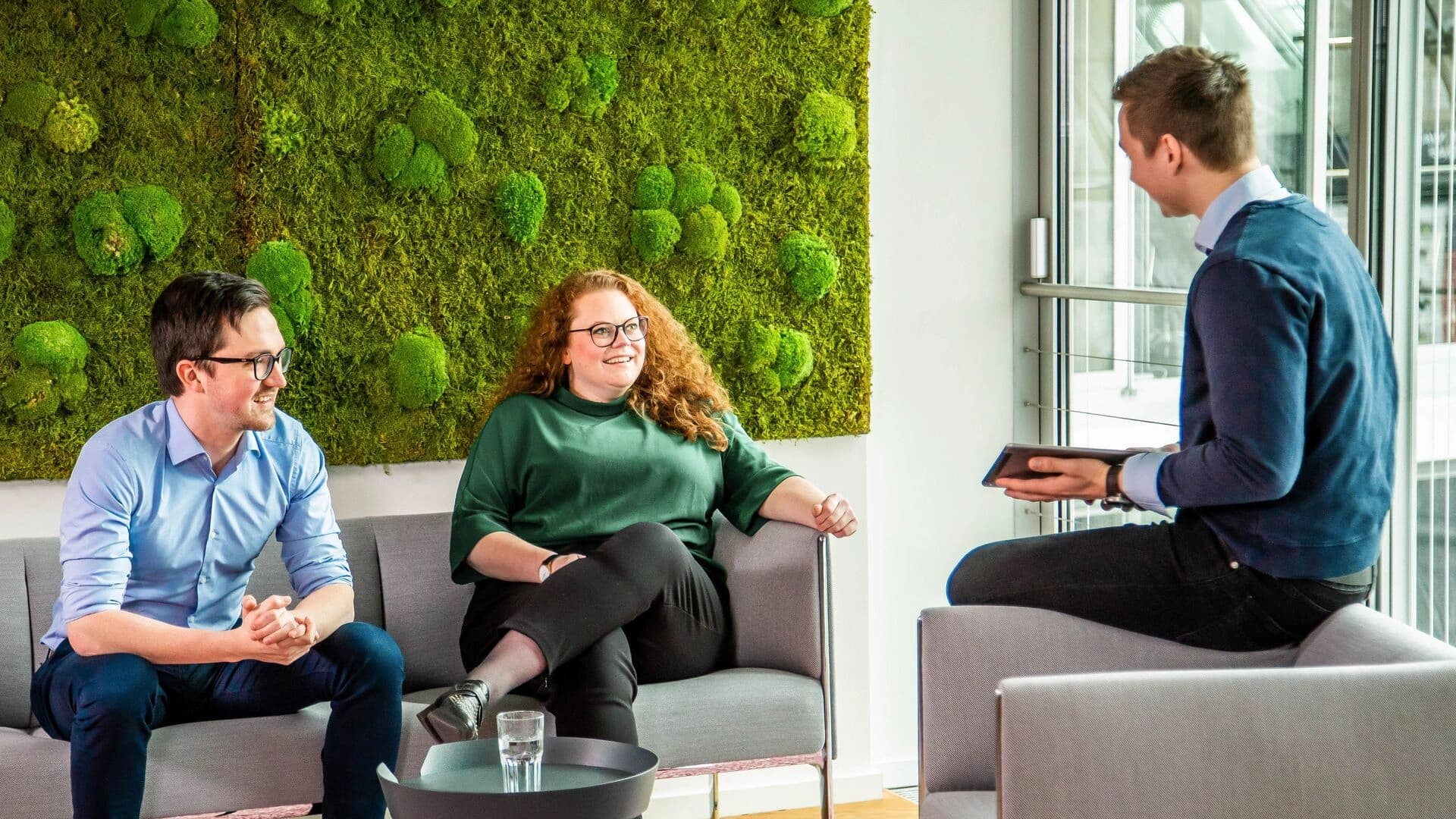 Three people are having a conversation in a modern Körber office lounge area, featuring a green moss wall and a large window in the background.