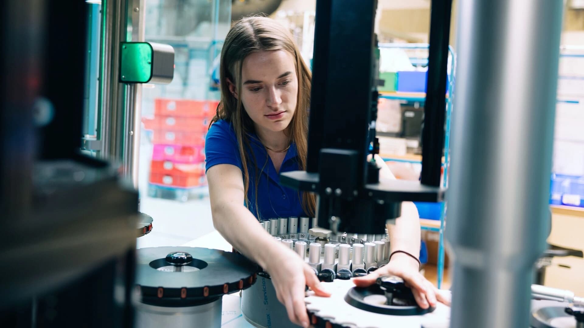 A student wearing a blue shirt operates machinery in an industrial or laboratory setting, carefully adjusting components on a circular metal device.